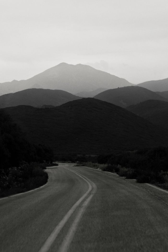 A dramatic black and white shot of a winding road leading to distant mountains under a moody sky.